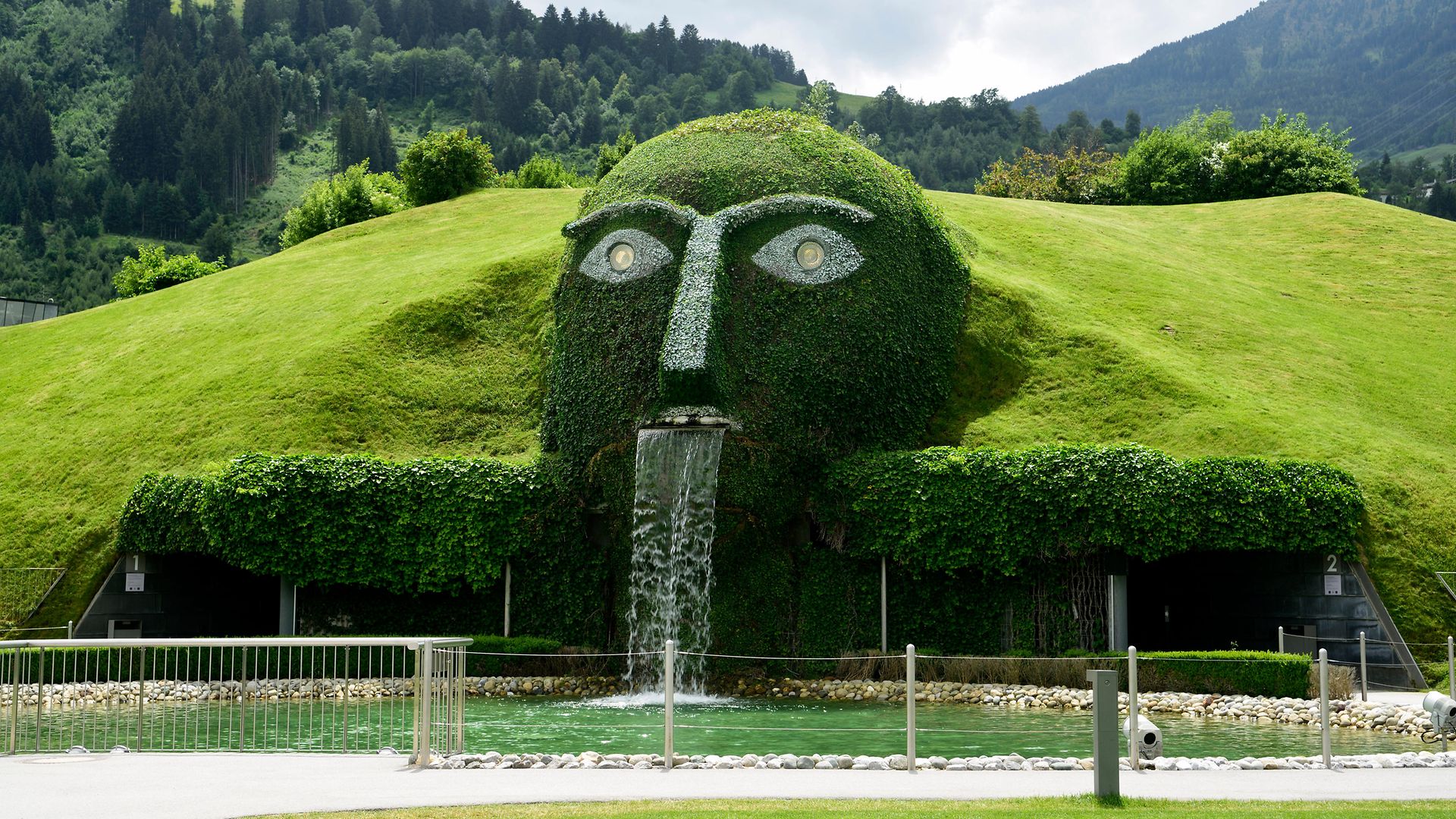 Springbrunnen mit Riesenkopf, der Wasser in einen Teich in den Swarowski Kristallwelten in Wattens spuckt, mit Vegetation.