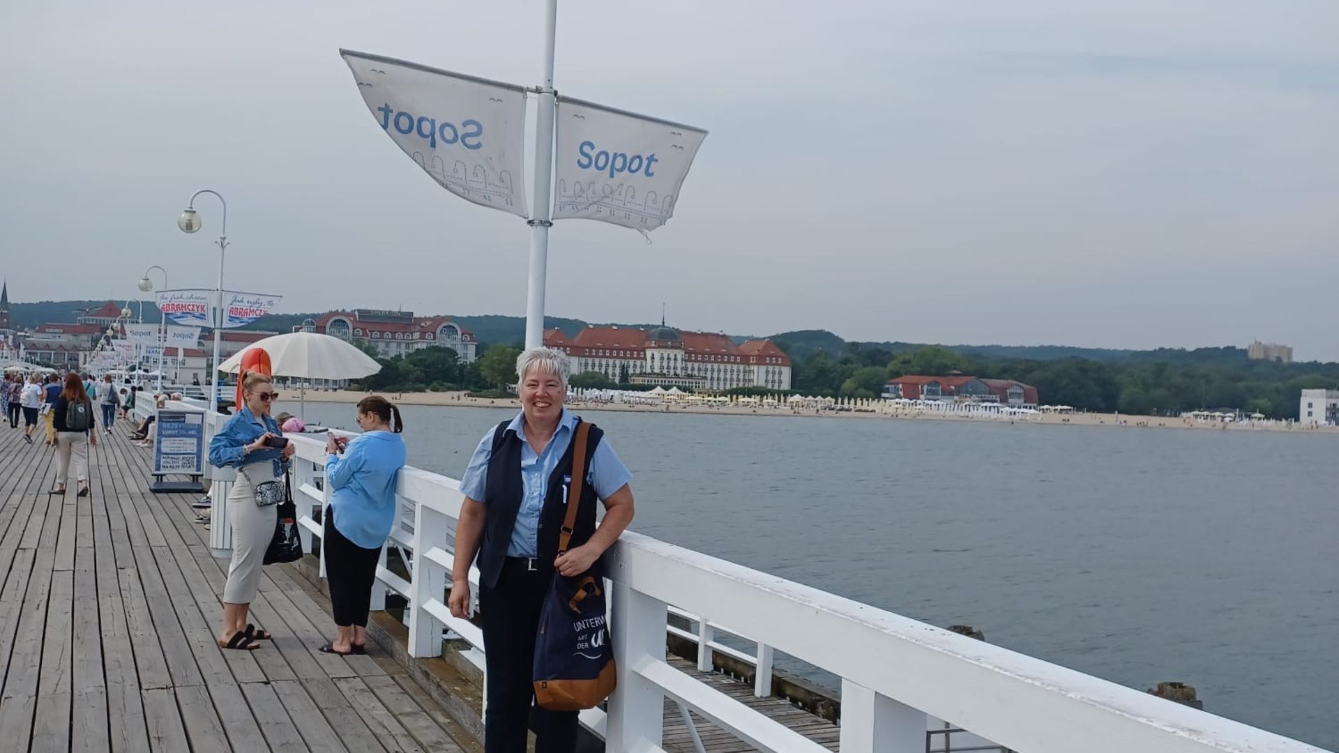 Frau steht vor einer Fahne auf der "Sopot" geschrieben steht. Hinter ihr ist ein Steg und Wasser